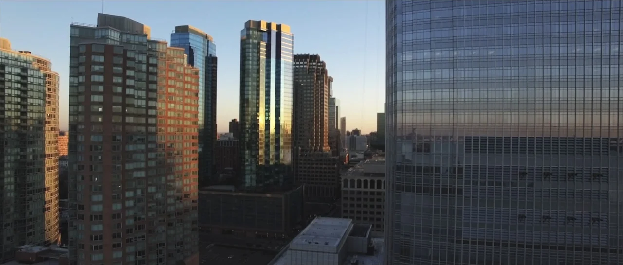 Square One (2019) movie still: establishing — A dense urban cluster of modern glass skyscrapers stands under a clear dusk sky. The buildings display…