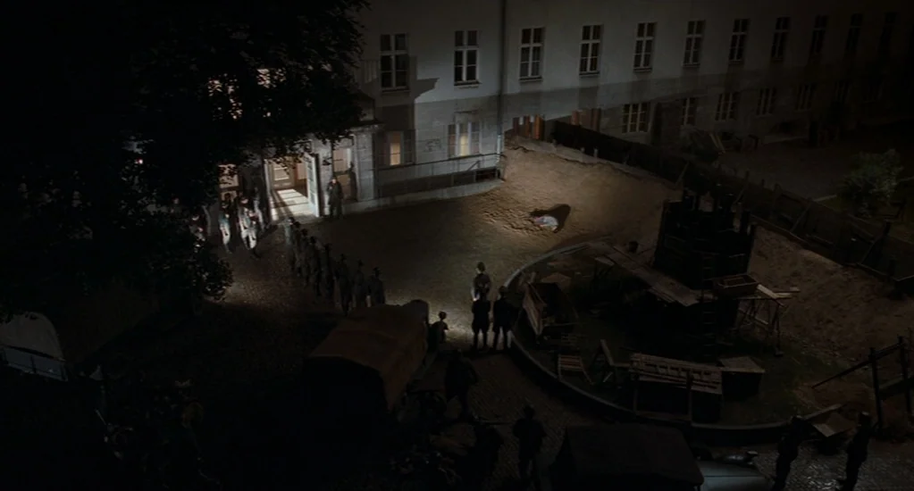 Valkyrie (2008) movie still: wide — A dark courtyard at night features a line of soldiers standing near a building. A single body lies on the dirt…