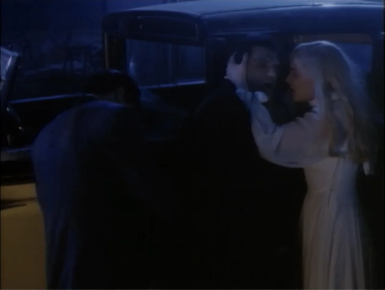 Cast a Deadly Spell (1991) movie still: medium — An old dark sedan sits at night with two people standing beside the door. A woman in a…