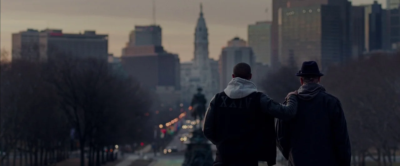 Creed (2015) movie still: wide — A public park path overlooks a distant city skyline with a tall clock tower. Two men stand together with their…