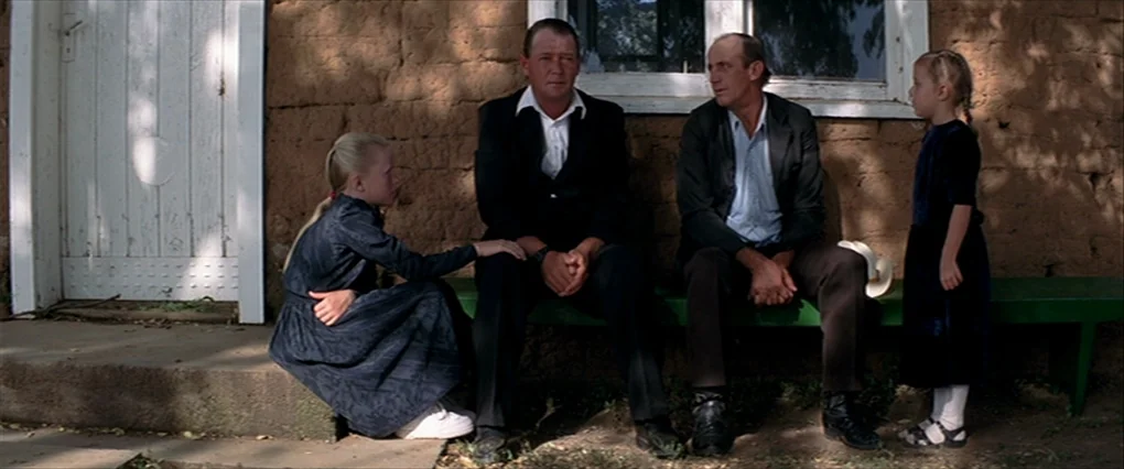 Silent Light (2007) movie still: fullbody — A group of people sits and stands against an unpainted adobe-style farmhouse wall. Two men occupy a…