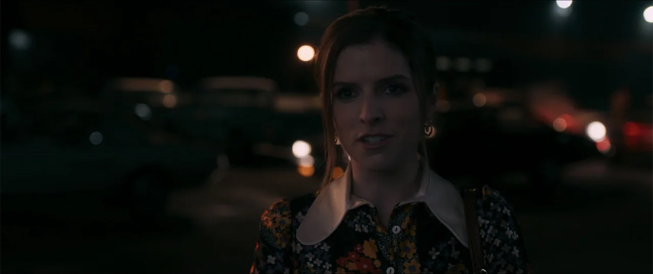 Woman of the Hour (2024) movie still: medium — An outdoor parking lot at night features blurred lights in the dark background. A woman in a floral…