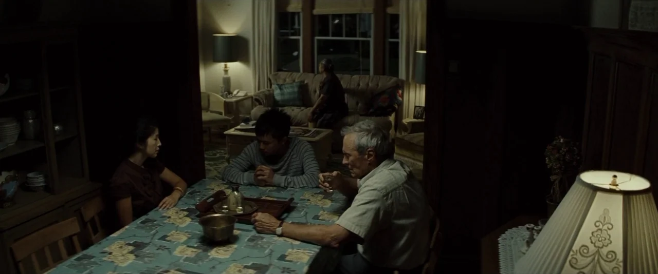 Gran Torino (2008) movie still: medium — A dimly lit dining area with dark wood furniture and patterned tablecloth. Three people sit around a table…