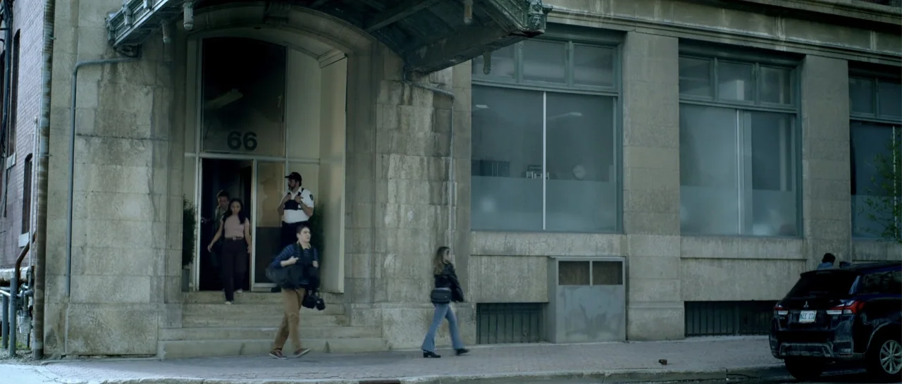Elevator Game (2023) movie still: wide — A stone building entrance features a large arched doorway and modern frosted glass windows. Three…