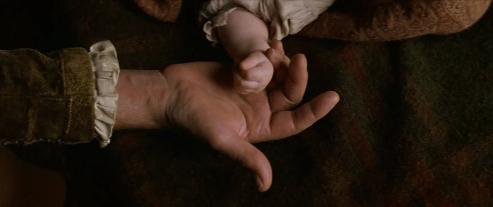 The New World (2005) movie still: closeup — A close-up view shows a fabric surface with textures of dark brown cloth. An adult hand lies palm up…