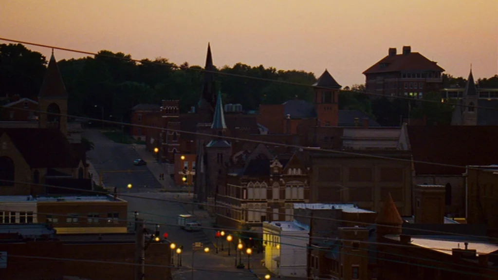 Sugar (2008) movie still: establishing — A quiet small-town street is flanked by low-rise brick buildings and church steeples. Power lines…