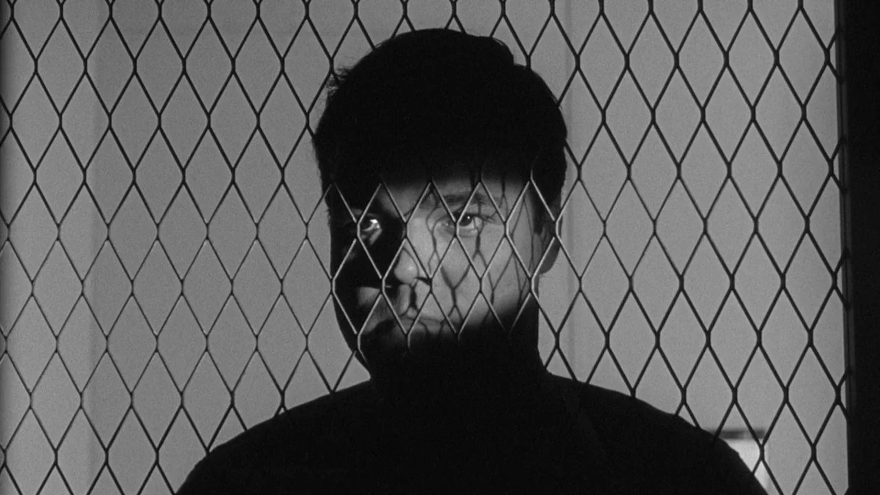 Shock Corridor (1963) movie still: closeup — A man stands behind a metal fence in a dark space. He looks toward the camera while the mesh creates a…