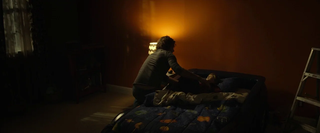 Before I Wake (2016) movie still: medium — A dimly lit room features orange walls and a child's bed with space-print blankets. A man leans over the…