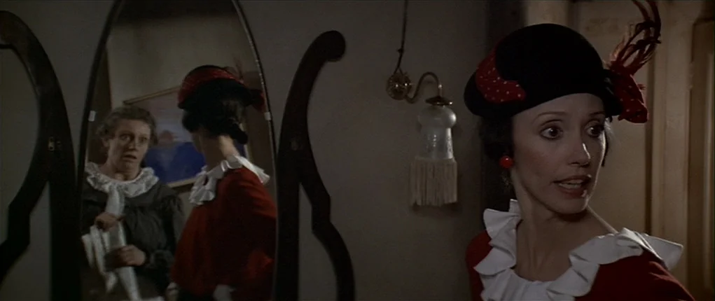 Popeye (1980) movie still: medium — A dark dressing room interior features a tall oval vanity mirror mounted on a wall. A woman in a red ruffled top…