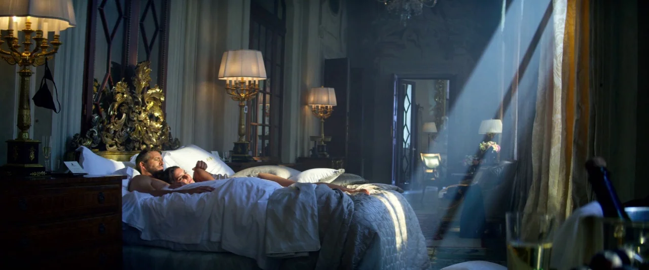 6 Underground (2019) movie still: wide — An opulent room features a large bed with ornate gold headboards and golden lamps. A man and a woman lie in…
