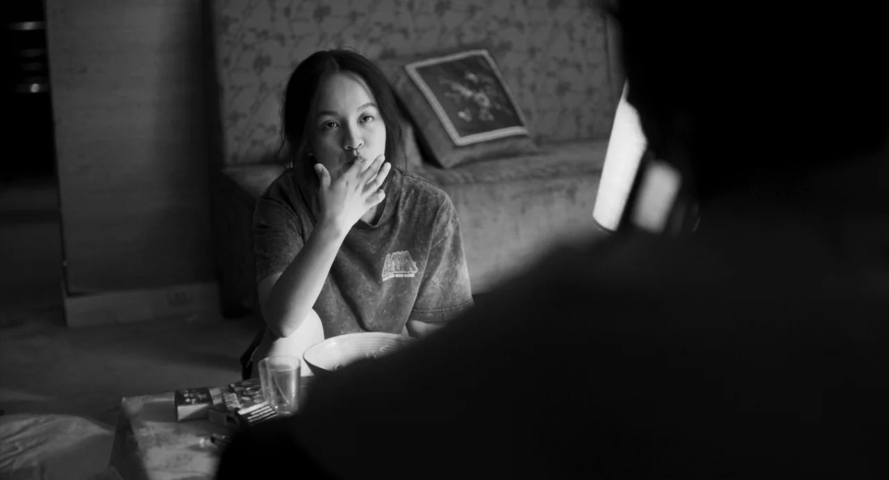 Paris, 13th District (2021) movie still: medium — A dark living room interior with a couch and small table in the background. A young woman sitting…