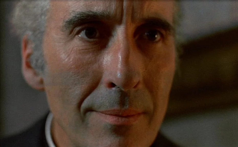 To The Devil a Daughter (1976) movie still: closeup — An elderly man is framed in a close-up shot inside a dark room. He stares at the camera with a…