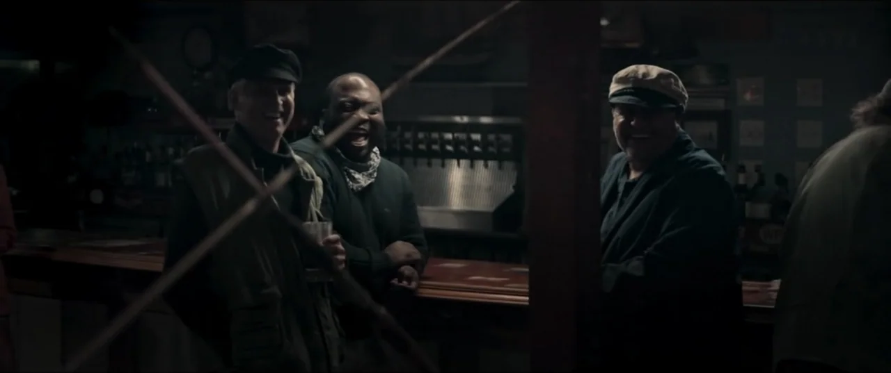 Offseason (2022) movie still: medium — A dimly lit bar interior features a wooden counter and shadowy background shelves. Three men stand behind the…