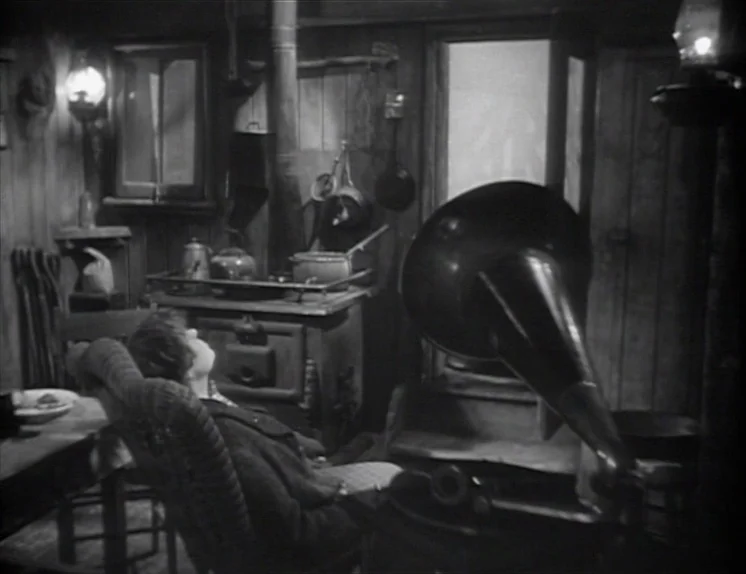 Anna Christie (1930) movie still: medium — A rustic wood-paneled room features a vintage wood-burning stove and a phonograph player. A lone…