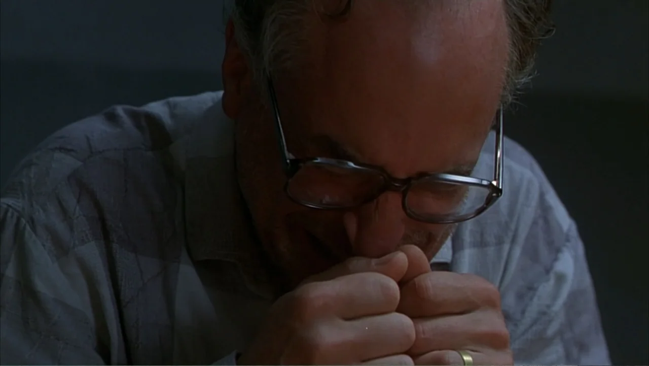 Citizen X (1995) movie still: closeup — A man sits in a dimly lit, nondescript interior space with a muted color palette. He sits with his eyes…