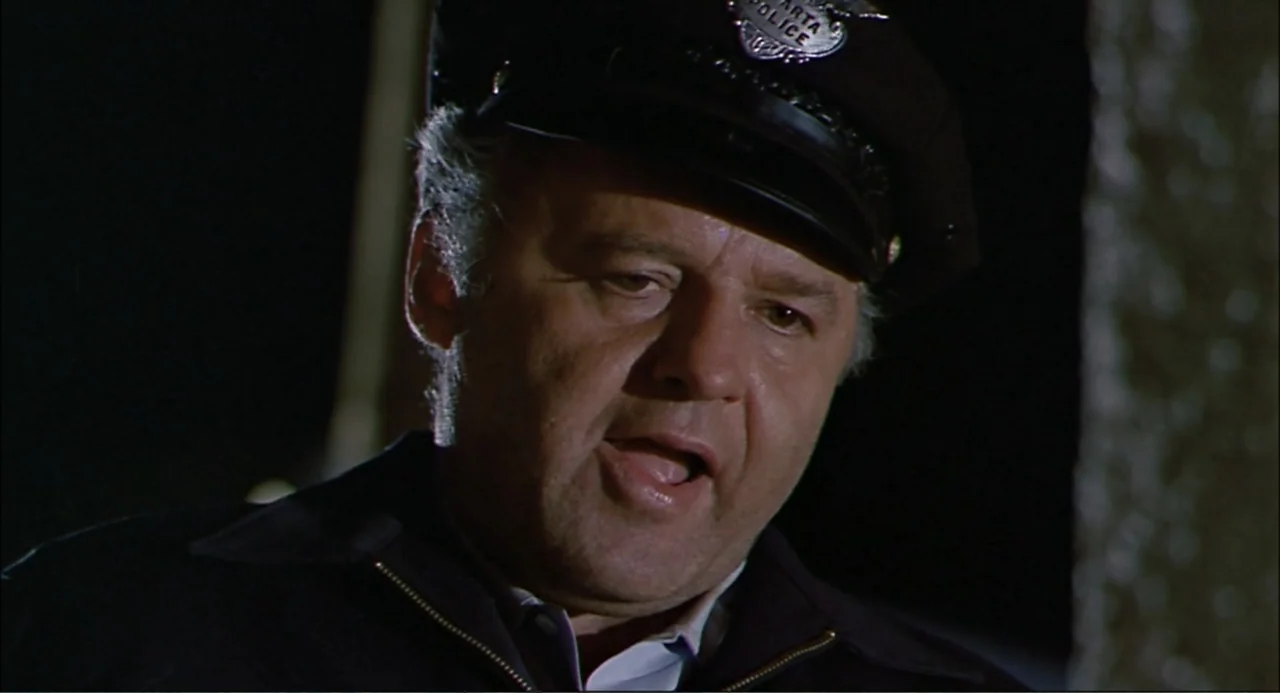In the Heat of the Night (1967) movie still: closeup — A man in a police uniform stands outdoors at night near a textured wall. The officer looks…