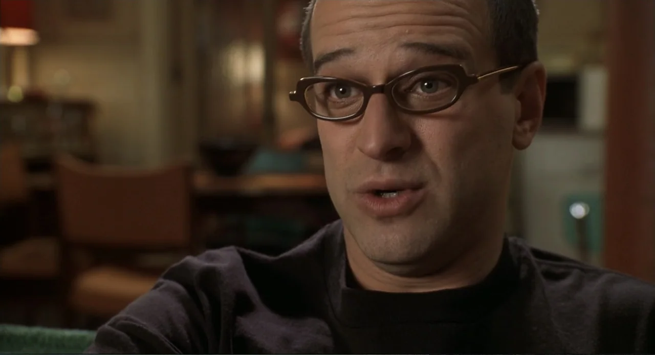 Little Nicky (2000) movie still: closeup — A dimly lit interior space contains a person in a darkened living room area. A man wearing glasses faces…