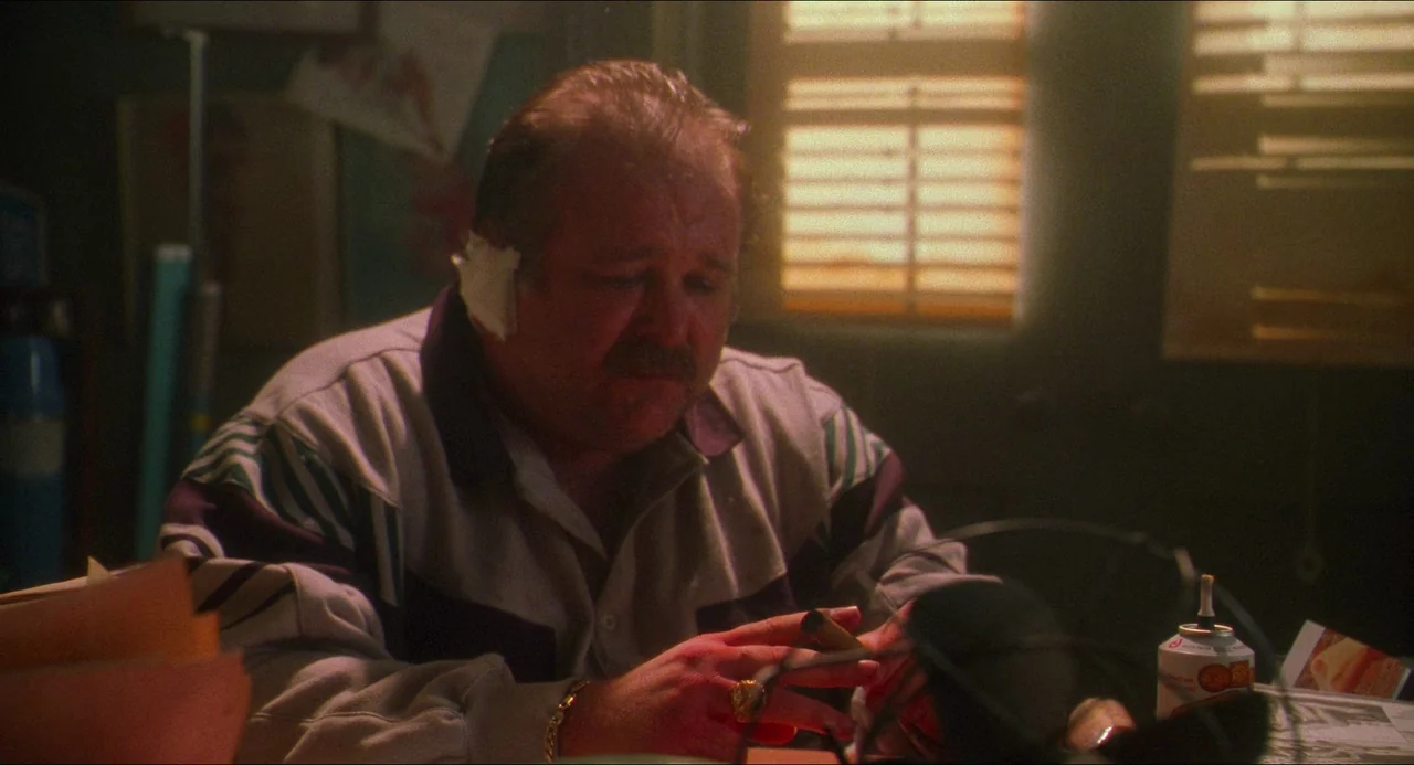 Hard Target (1993) movie still: medium — An office space with venetian blinds on the window filtering daylight. A man with a bandaged ear sits at a…