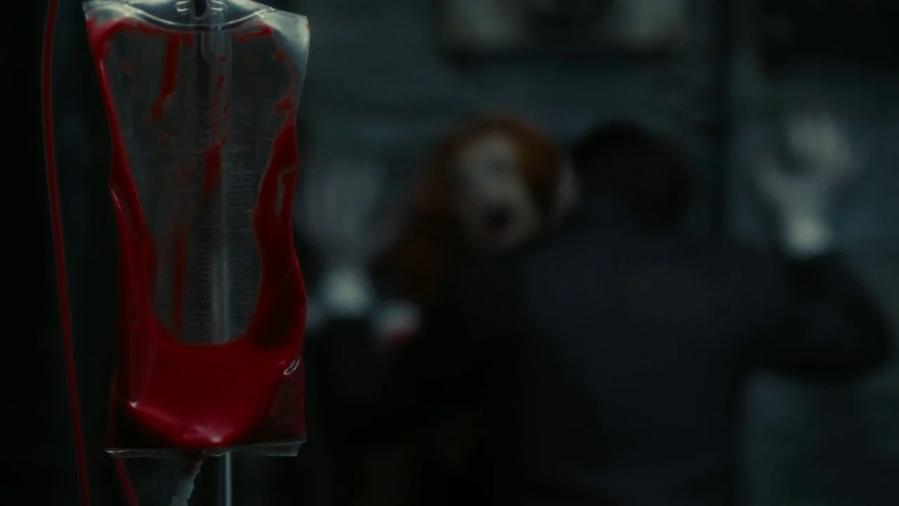 Dark Shadows (2012) movie still: closeup — A medical intravenous bag filled with bright red blood hangs in the sharp foreground. Two blurred figures…