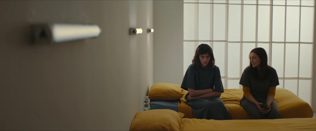 Double Blind (2023) movie still: medium — A spare, clinical room with white walls, two single beds, and a gridded window. Two women sit side-by-side…