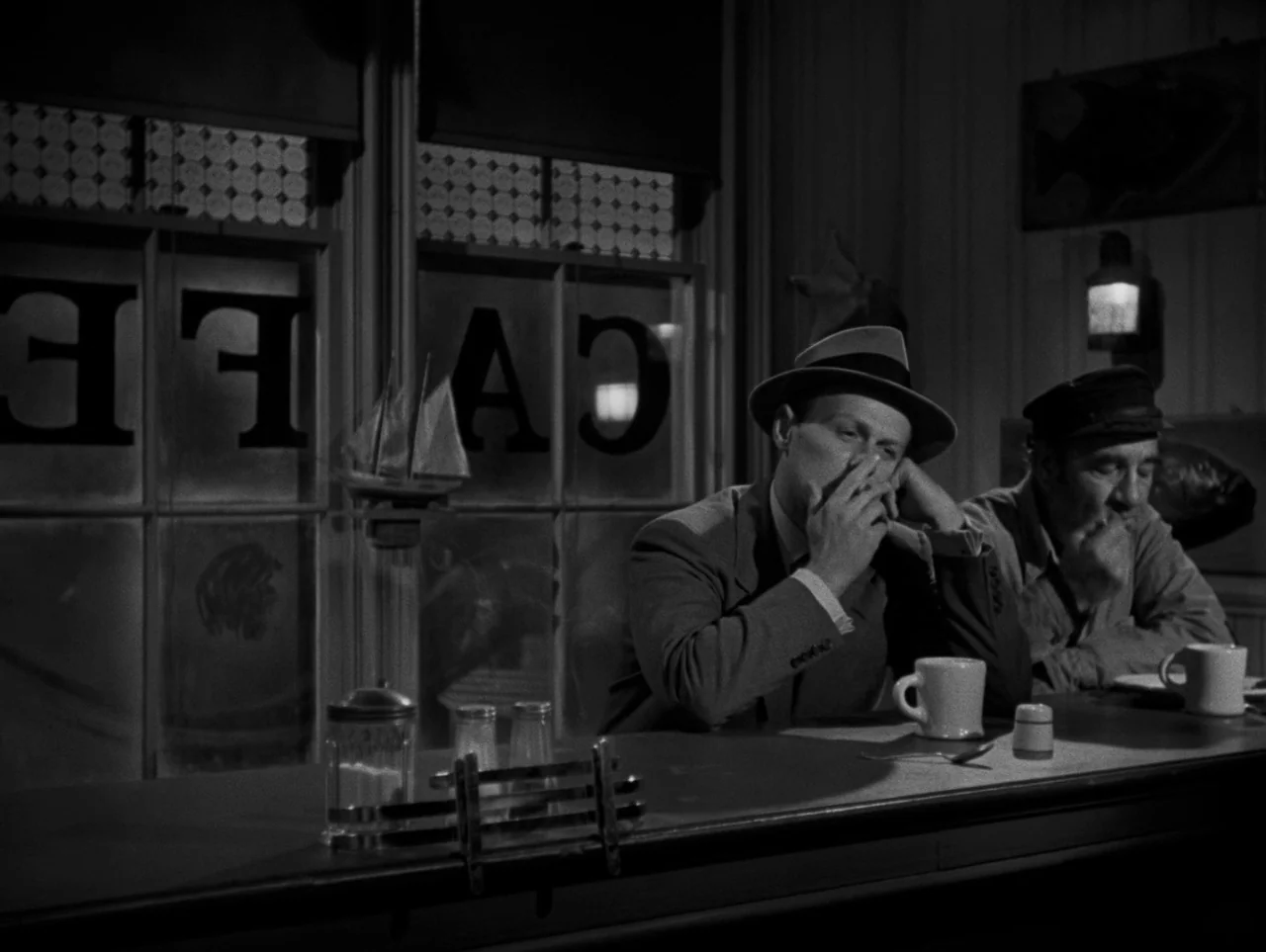 Pickup on South Street (1953) movie still: medium — A dimly lit diner with a glass window displaying backward lettering reading CAFE. Two men sit at…