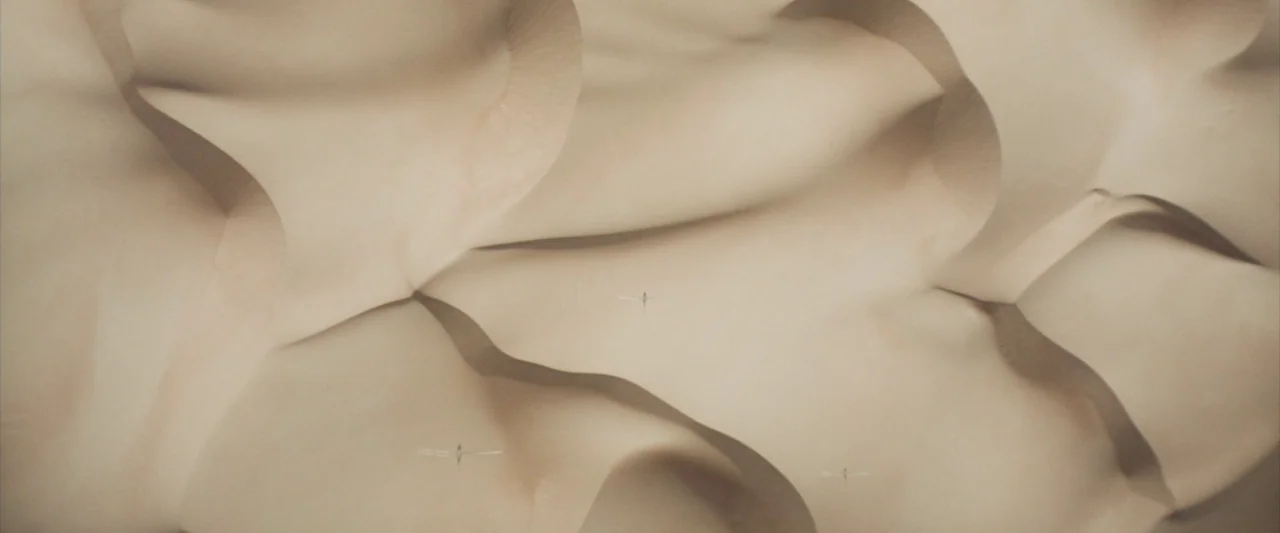 Dune (2021) movie still: establishing — Expansive desert dunes stretch across the frame with smooth, undulating surfaces of sand. Three small…