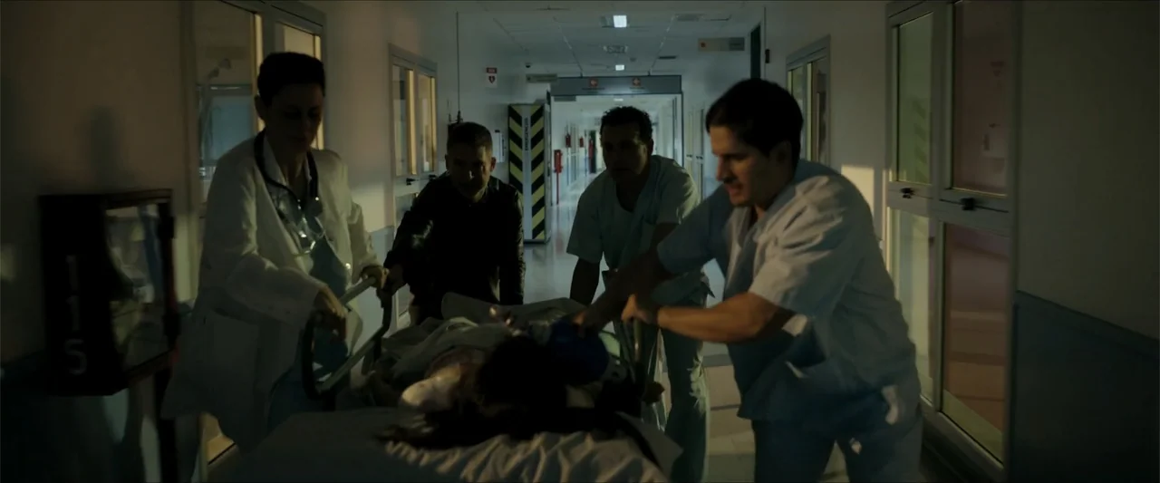 Checkmate (2015) movie still: medium — A long hospital corridor with glass doors and white walls serves as the backdrop. Medical staff and a person…