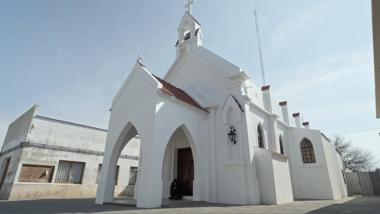 Saturdays Disorders (2022) movie still: wide — A small white church with red tile roof accents sits under a clear sky. A person sits quietly in the…