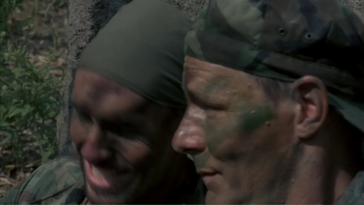 Behind Enemy Lines (2001) movie still: closeup — A dense outdoor location with green vegetation and a textured gray stone wall backdrop. Two…