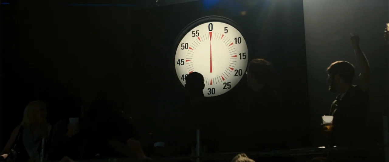 Now You See Me (2013) movie still: medium — A dark, low-light interior space features a large circular timer mounted on a wall. Several people stand…