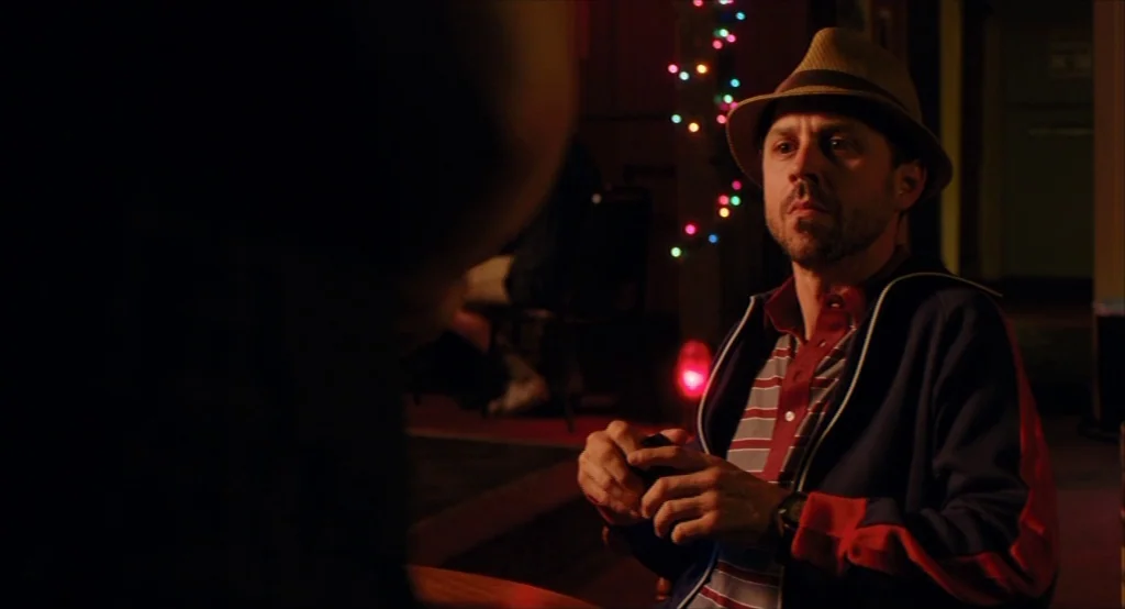 Results (2015) movie still: medium — A dimly lit room with blurred multi-colored decorative string lights in the background. A man wearing a fedora…