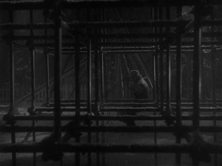 Ikiru (1952) movie still: medium — A dark, wet playground at night features a large, geometric metal jungle gym. A man sits motionless inside the…