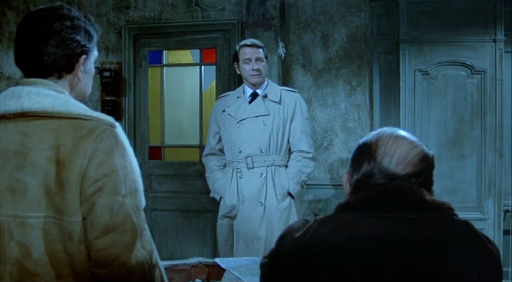 Un Flic (1972) movie still: medium — A dimly lit room features walls with textured, peeling wallpaper and a stained-glass door. A man in a beige…