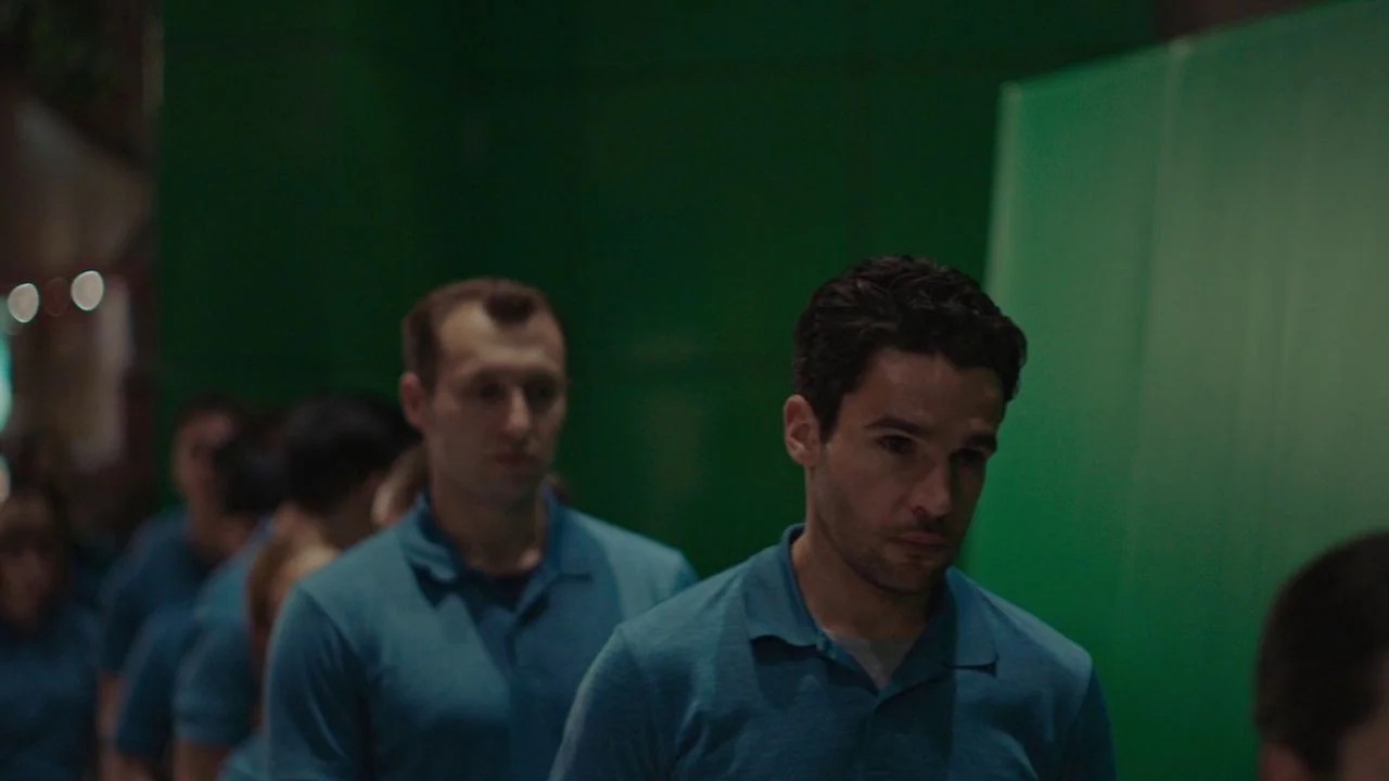 Possessor (2020) movie still: medium — A group of people wearing blue polo shirts stand before a large green screen. The man in the foreground moves…