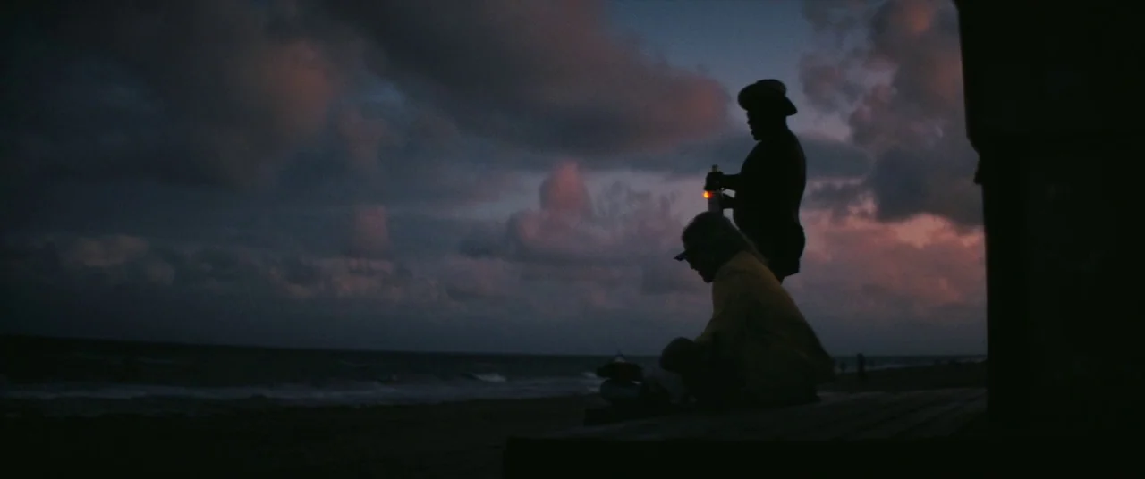 The Beach Bum (2019) movie still: wide — A dark wooden platform faces a calm ocean under a cloudy sunset sky. Two figures sit in silhouette on the…