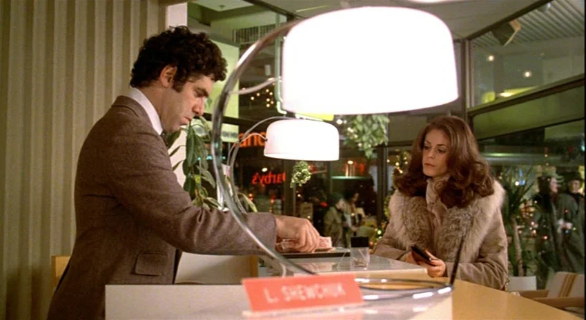 The Silent Partner (1978) movie still: medium — An office reception area featuring a desk with a white lamp and a sign. A man in a brown suit…