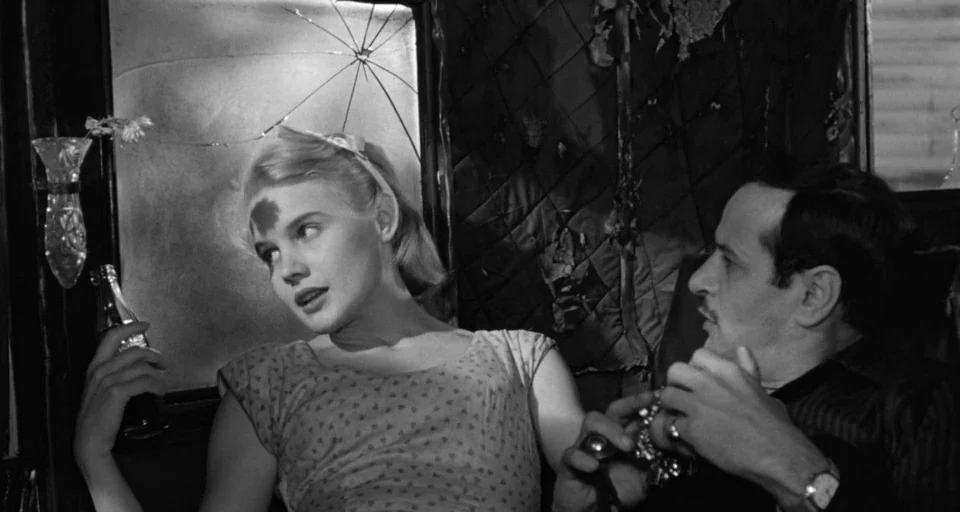 Baby Doll (1956) movie still: medium — A dark train compartment features a cracked windowpane and torn upholstery. A blonde woman sits beside a man…