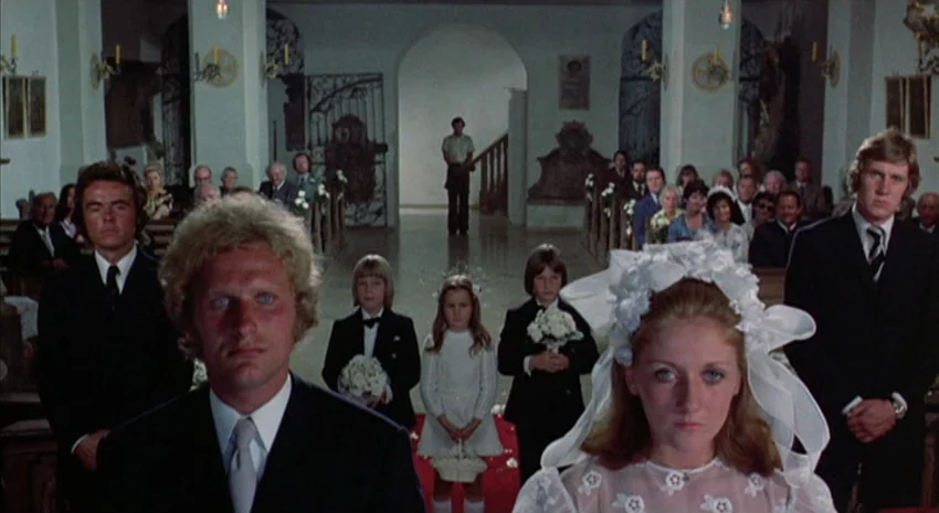 The Passenger (1975) movie still: medium — A white stone church interior with arched columns and wooden pews filled with guests. A wedding party…