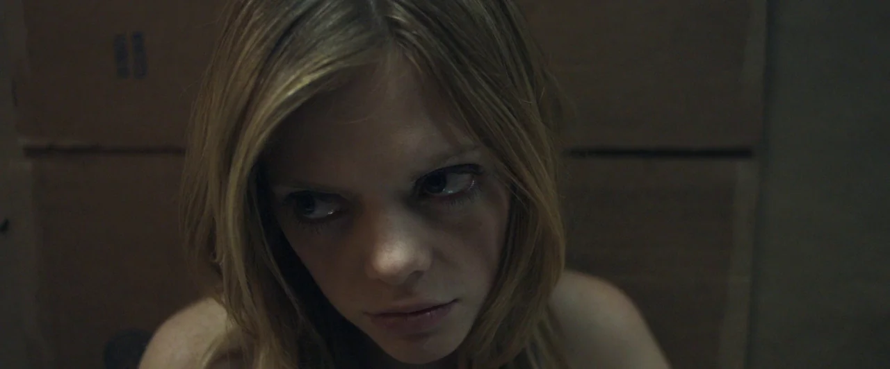 Compliance (2012) movie still: closeup — A dimly lit interior space with plain cardboard surfaces forming the backdrop. A woman with blonde hair…