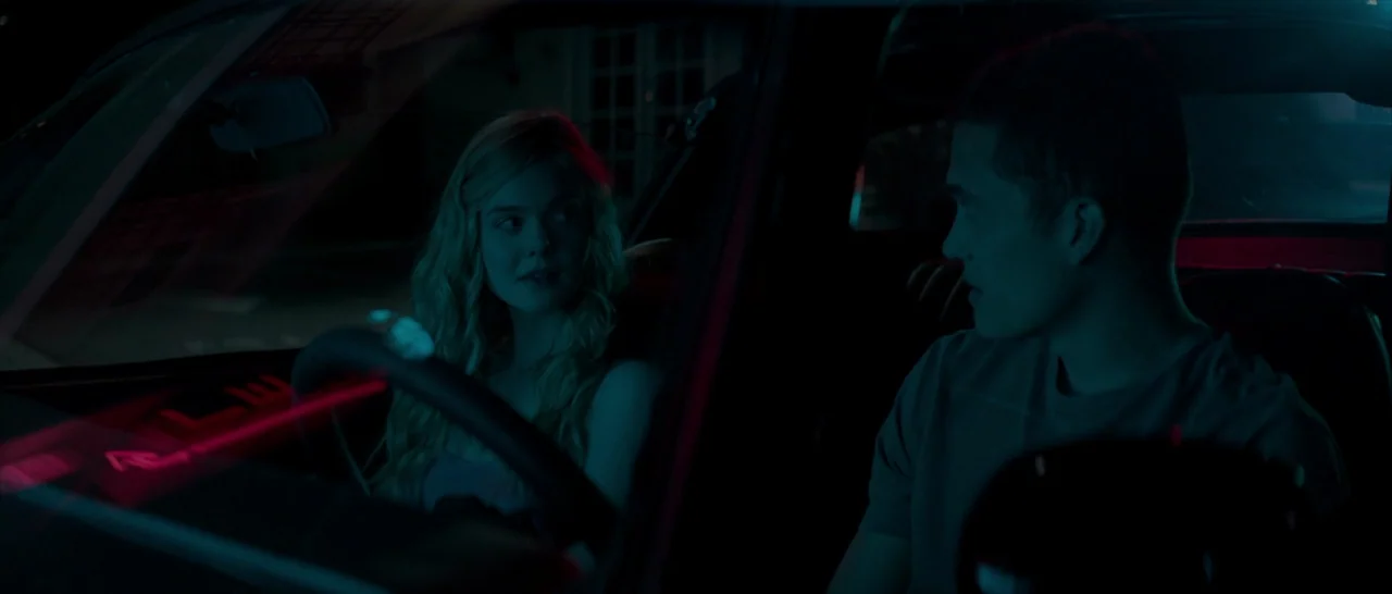 The Neon Demon (2016) movie still: medium — The interior of a parked car at night is illuminated by vibrant neon dashboard lighting. A young man and…