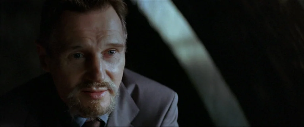 Batman Begins (2005) movie still: closeup — A dimly lit indoor space with blurred architectural curves in the background. A man in a suit looks…