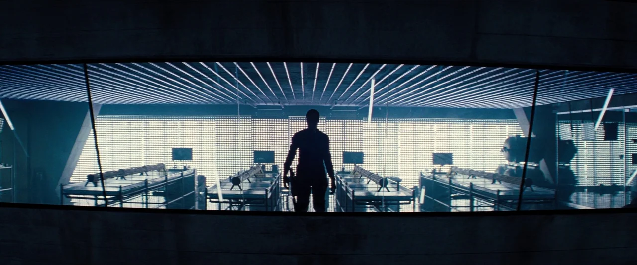 No Time To Die (2021) movie still: wide — A dark laboratory features a long row of stainless steel tables with linear objects. A lone figure stands…