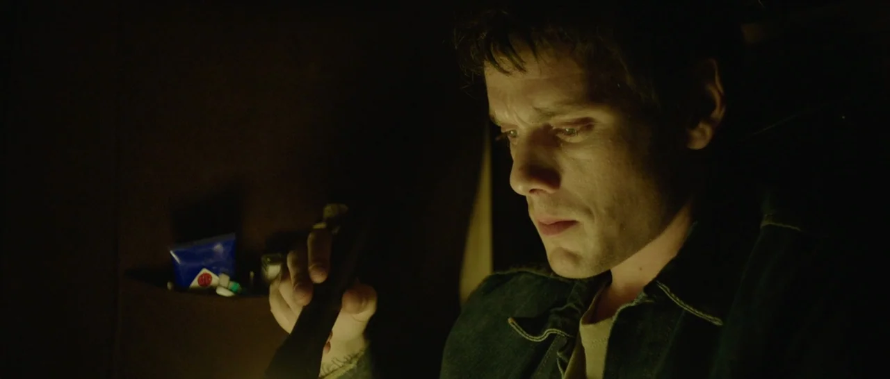 Green Room (2015) movie still: closeup — The interior of a dark vehicle at night with minimal illumination. A young man looks down at a handheld…
