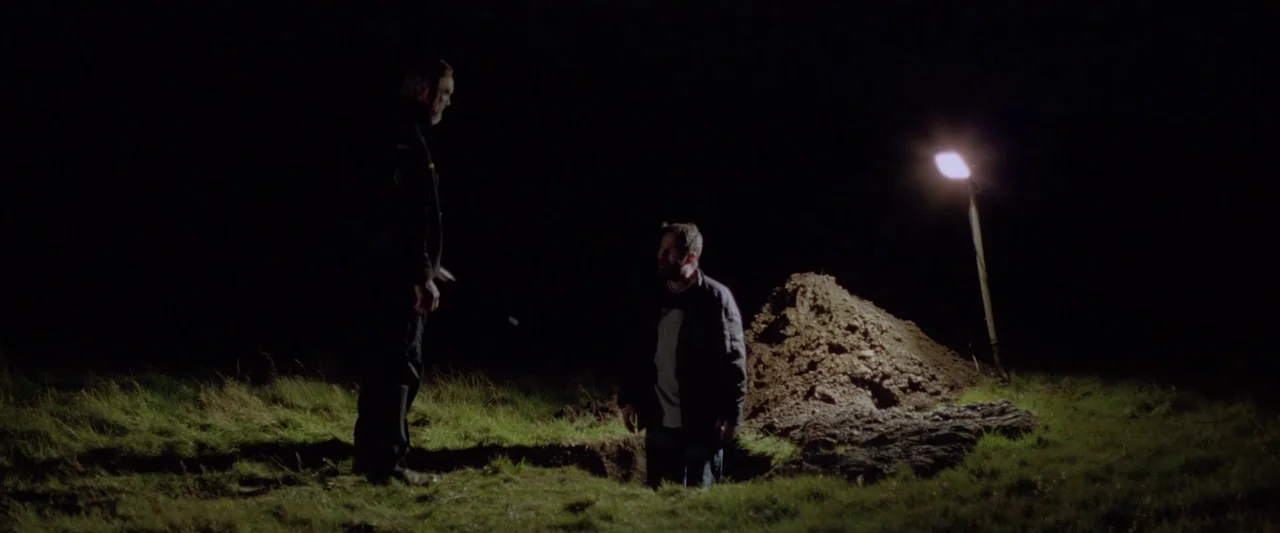 A White, White Day (2019) movie still: wide — An open pit in a grassy field is illuminated by a portable work light. Two men face each other near a…