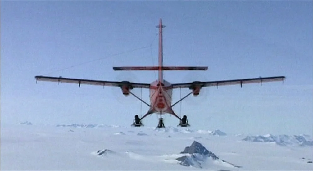 9 Songs (2004) movie still: wide — A red and white airplane flies over a vast, flat expanse of Arctic snow. Snow-covered peaks and jagged rock…