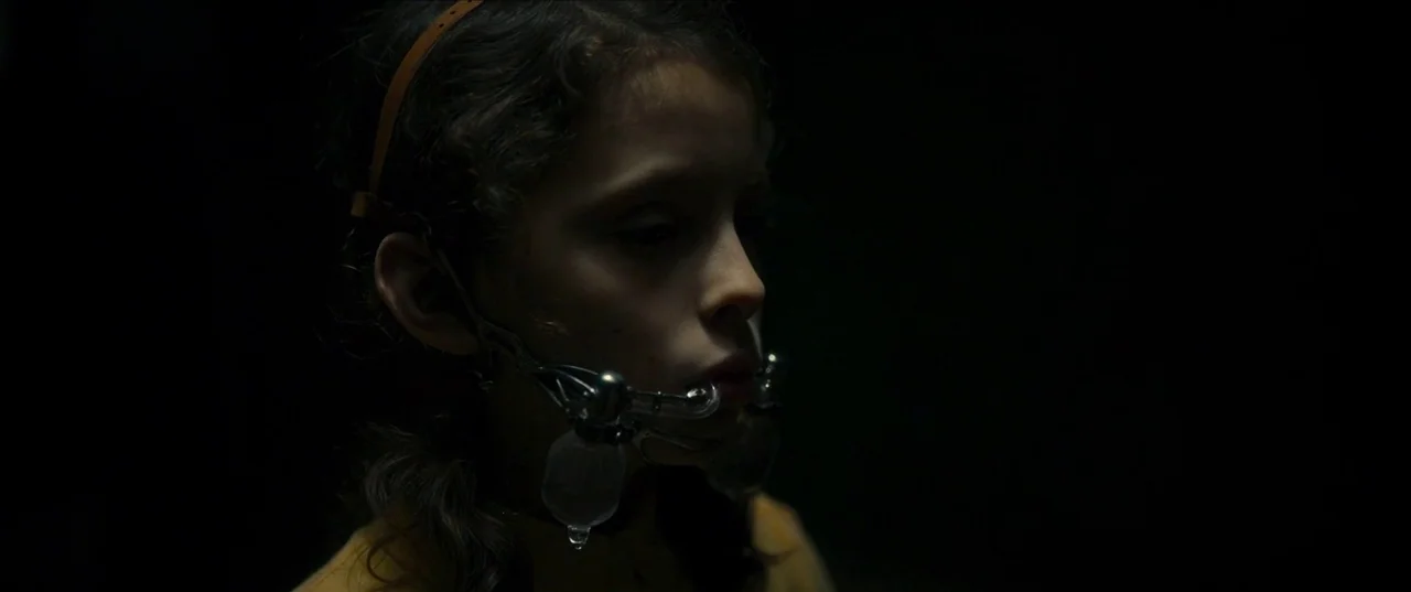 Earwig (2022) movie still: closeup — A young child is centered against a pitch-black background with no visible setting. The child wears a metal…