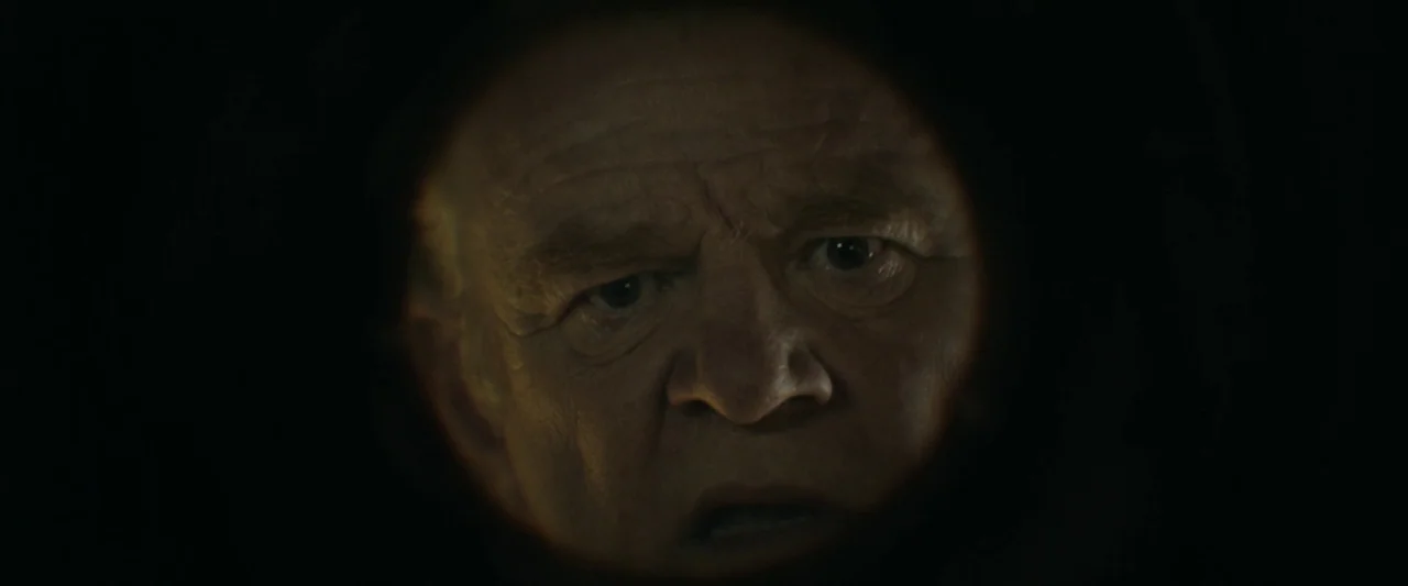 The Banshees of Inisherin (2022) movie still: closeup — A dark space seen through a circular peephole lens on a door. An older man stares intently…