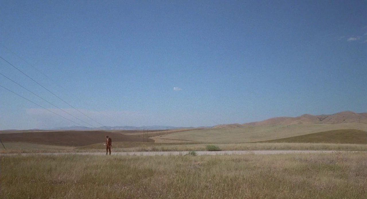 Napoleon Dynamite (2004) movie still: wide — A vast, open rural landscape with rolling tan hills and a clear blue sky. A lone man in a brown suit…