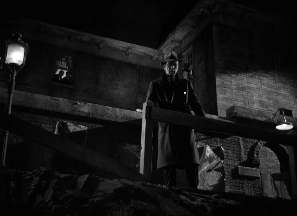 The Maltese Falcon (1941) movie still: fullbody — A dark brick building facade looms above a wooden railing at night. A man in a trench coat and…