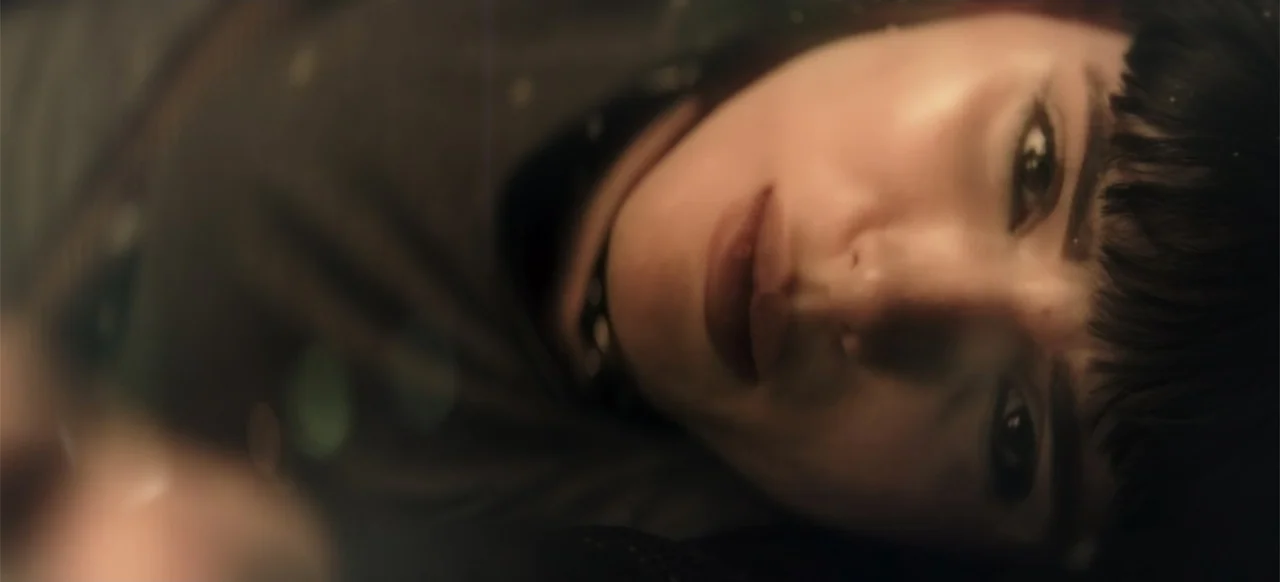 I Still See You (2018) movie still: closeup — A dimly lit interior space with shadows obscuring the surroundings. A young woman with short dark hair…